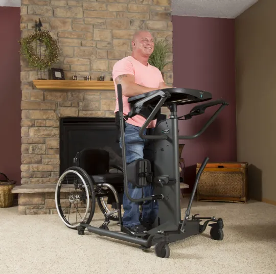 Man standing in a sit-to-stand standing frame with a tray, his wheelchair behind him in a living room